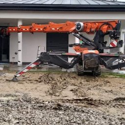 ARCHIFORMA ARCHITEKCI GROUP SPÓŁKA Z OGRANICZONĄ ODPOWIEDZIALNOŚCIĄ - Pomarańczowy żuraw pająk ustawiony na błotnistym terenie przed nowoczesnym, parterowym domem z dużymi oknami i szarą dachówką.