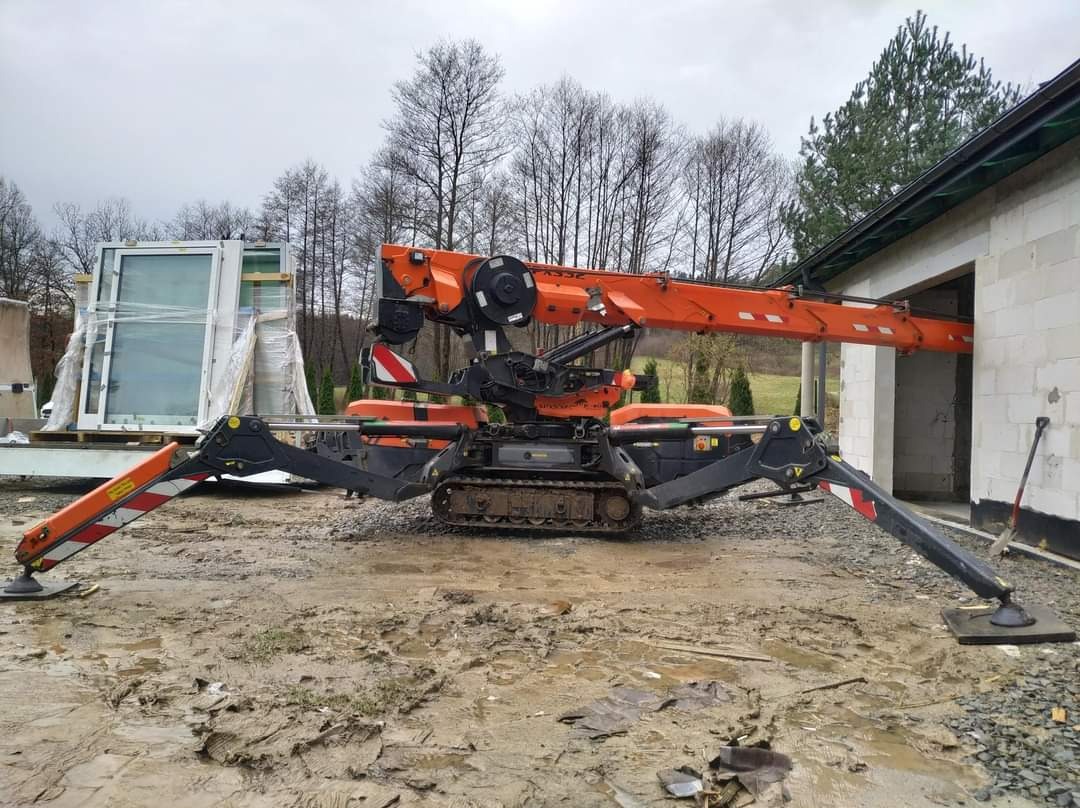 Pająkowa mini żuraw gąsienicowy z wysuniętym ramieniem w stronę otworu w surowym budynku z bloczków, na błotnistym placu budowy, w tle okna do montażu.