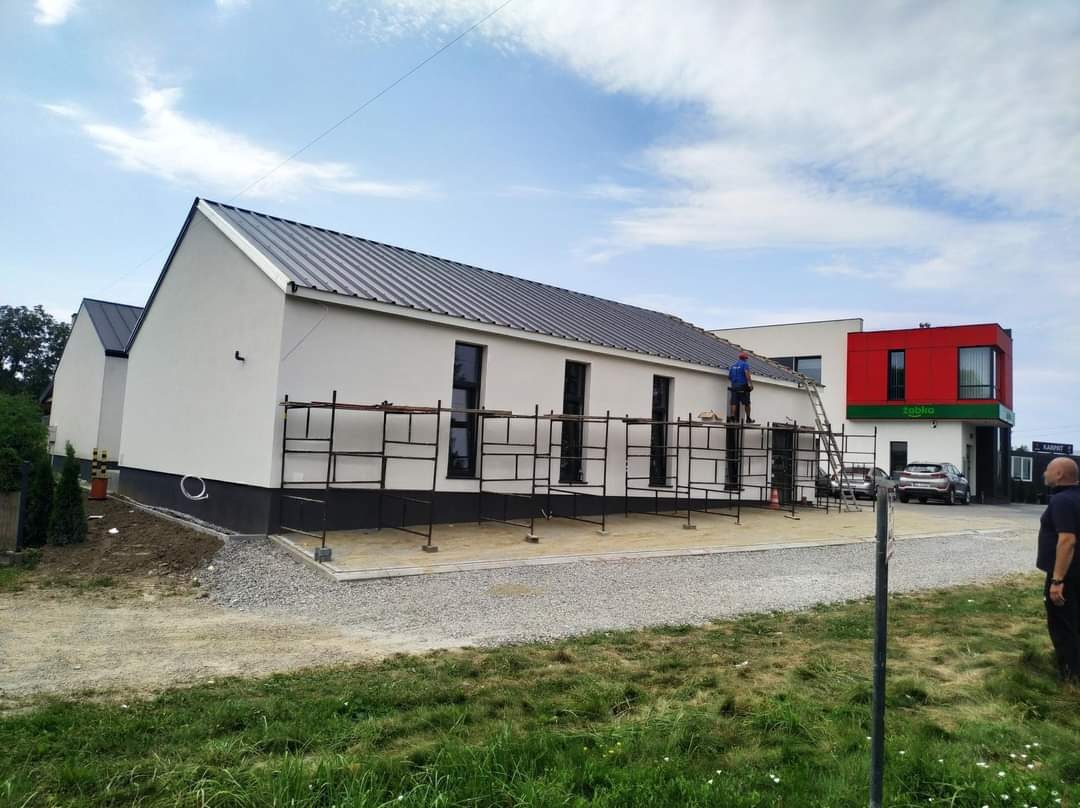 Elewacja budynku usługowego z rusztowaniem, pracownik na dachu w trakcie prac wykończeniowych, widoczny fragment sklepu Żabka w tle.