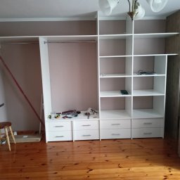Mariusz Mroziński - Montaż szafy na wymiar w Słupi: widoczne elementy szafy z szufladami i półkami, narzędzia na blacie, drewniana podłoga i stołek obok ściany.