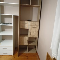 Mariusz Mroziński - Fragment pokoju z drewnianą podłogą, na której stoją dwa regały: biały z szufladami i otwartymi półkami oraz drewniany regał z szufladami, szafką i otwartymi półkami, w tle beżowa ściana i biała...