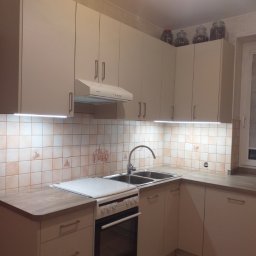 Mariusz Mroziński - Jasna kuchnia z kremowymi szafkami, białym okapem i płytą grzewczą; na górnych szafkach słoiki z przyprawami, płytki ceramiczne z motywami rustykalnymi.