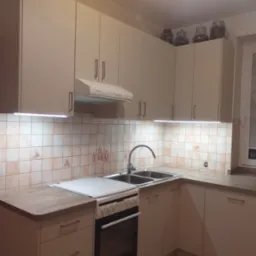 Jasna kuchnia z kremowymi szafkami, białym okapem i płytą grzewczą; na górnych szafkach słoiki z przyprawami, płytki ceramiczne z motywami rustykalnymi.