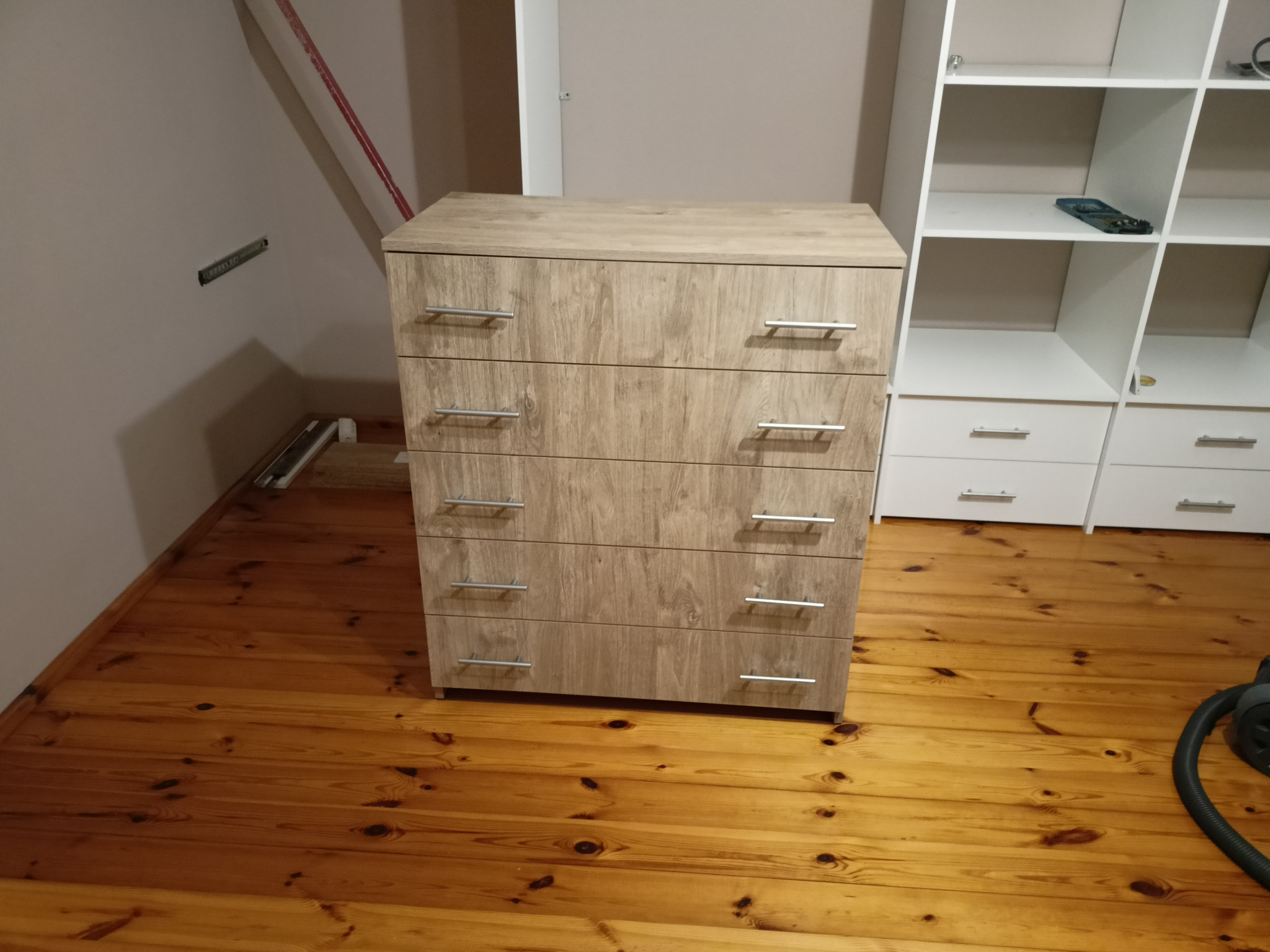 Jasna komoda z czterema szufladami z metalowymi uchwytami, stojąca na drewnianej podłodze, obok biały regał z półkami.