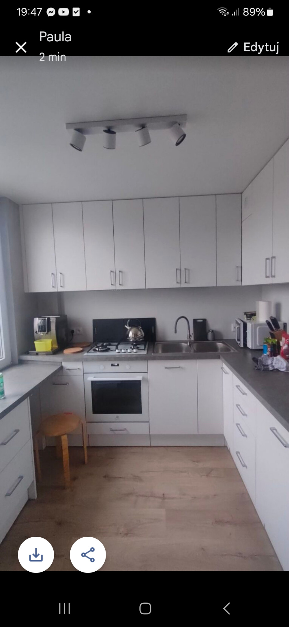 Jasna kuchnia w kształcie litery U z białymi szafkami, drewnianą podłogą i szarym blatem, oświetlona reflektorami sufitowymi, z widocznym czajnikiem na kuchence.