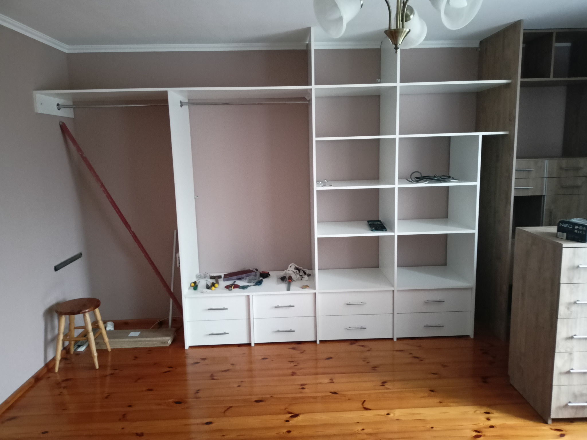 Montaż szafy na wymiar w Słupi: widoczne elementy szafy z szufladami i półkami, narzędzia na blacie, drewniana podłoga i stołek obok ściany.