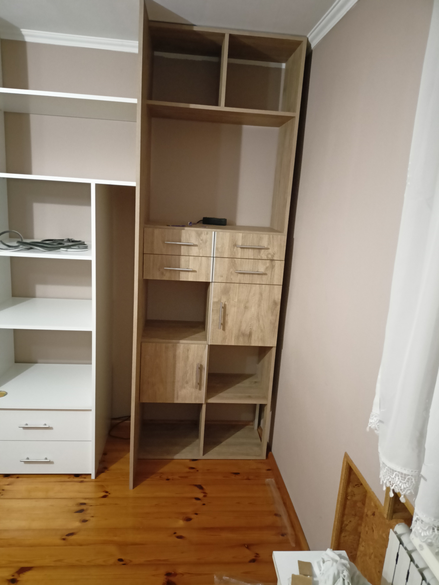 Fragment pokoju z drewnianą podłogą, na której stoją dwa regały: biały z szufladami i otwartymi półkami oraz drewniany regał z szufladami, szafką i otwartymi półkami, w tle beżowa ściana i biała...