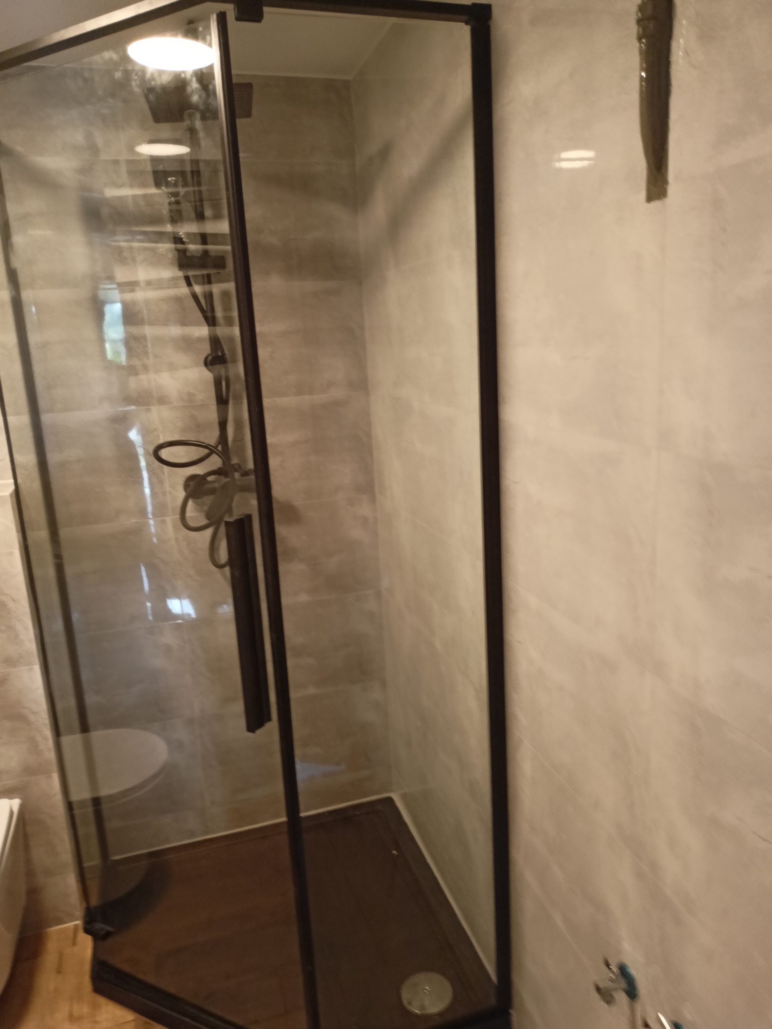 Nowoczesna kabina prysznicowa z czarnymi profilami i deszczownicą, wykończona płytkami imitującymi kamień, widoczna część toalety obok.