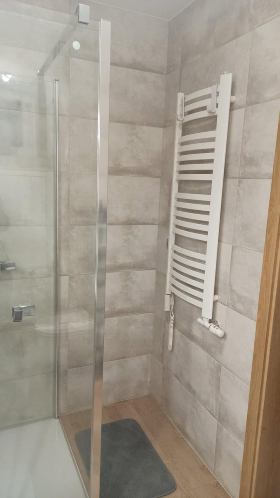 Nowoczesna kabina prysznicowa ze szklanymi drzwiami i chromowanymi okuciami, grzejnik drabinkowy w kolorze białym na ścianie wyłożonej płytkami w odcieniach szarości, szara wycieraczka pod prysznicem.