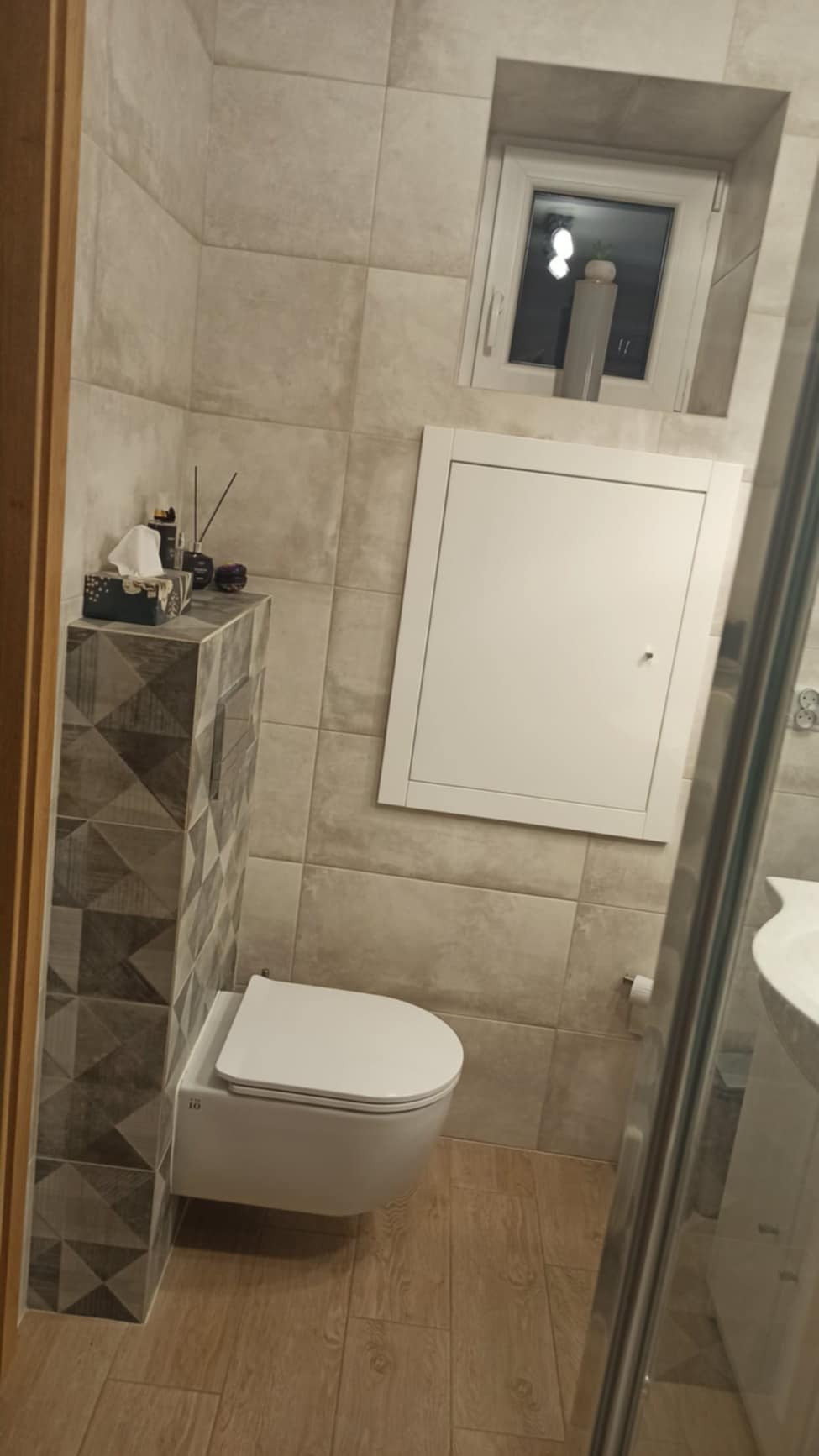 Nowoczesna toaleta podwieszana w łazience wykończonej płytkami w odcieniach szarości i beżu, z drewnopodobną podłogą i akcesoriami dekoracyjnymi na zabudowie stelaża.