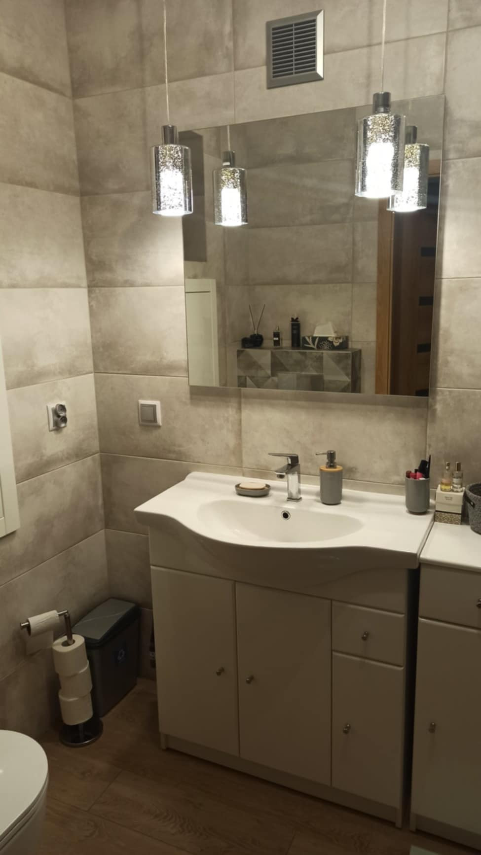 Umywalka z szafką w łazience z beżowymi płytkami, oświetlona trzema wiszącymi lampami z efektem brokatu, z widocznym koszem na śmieci i zapasem papieru toaletowego