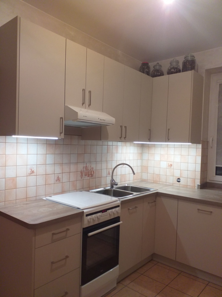 Jasna kuchnia z kremowymi szafkami, białym okapem i płytą grzewczą; na górnych szafkach słoiki z przyprawami, płytki ceramiczne z motywami rustykalnymi.