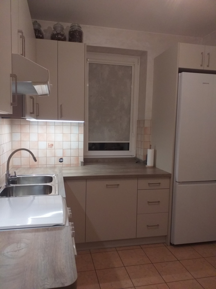 Jasna kuchnia z beżowymi szafkami, dwukomorowym zlewem, białym blatem i brązowymi płytkami podłogowymi, z trzema słoikami na górnych szafkach.