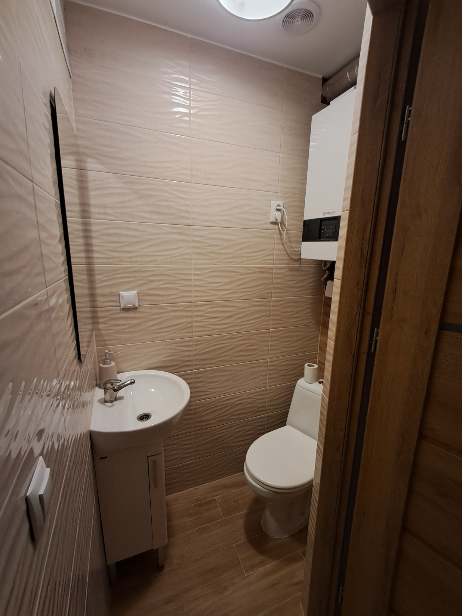 Wykończona łazienka z beżowymi płytkami o falistej strukturze, wiszącą umywalką z szafką, toaletą i piecem gazowym Buderus na ścianie.