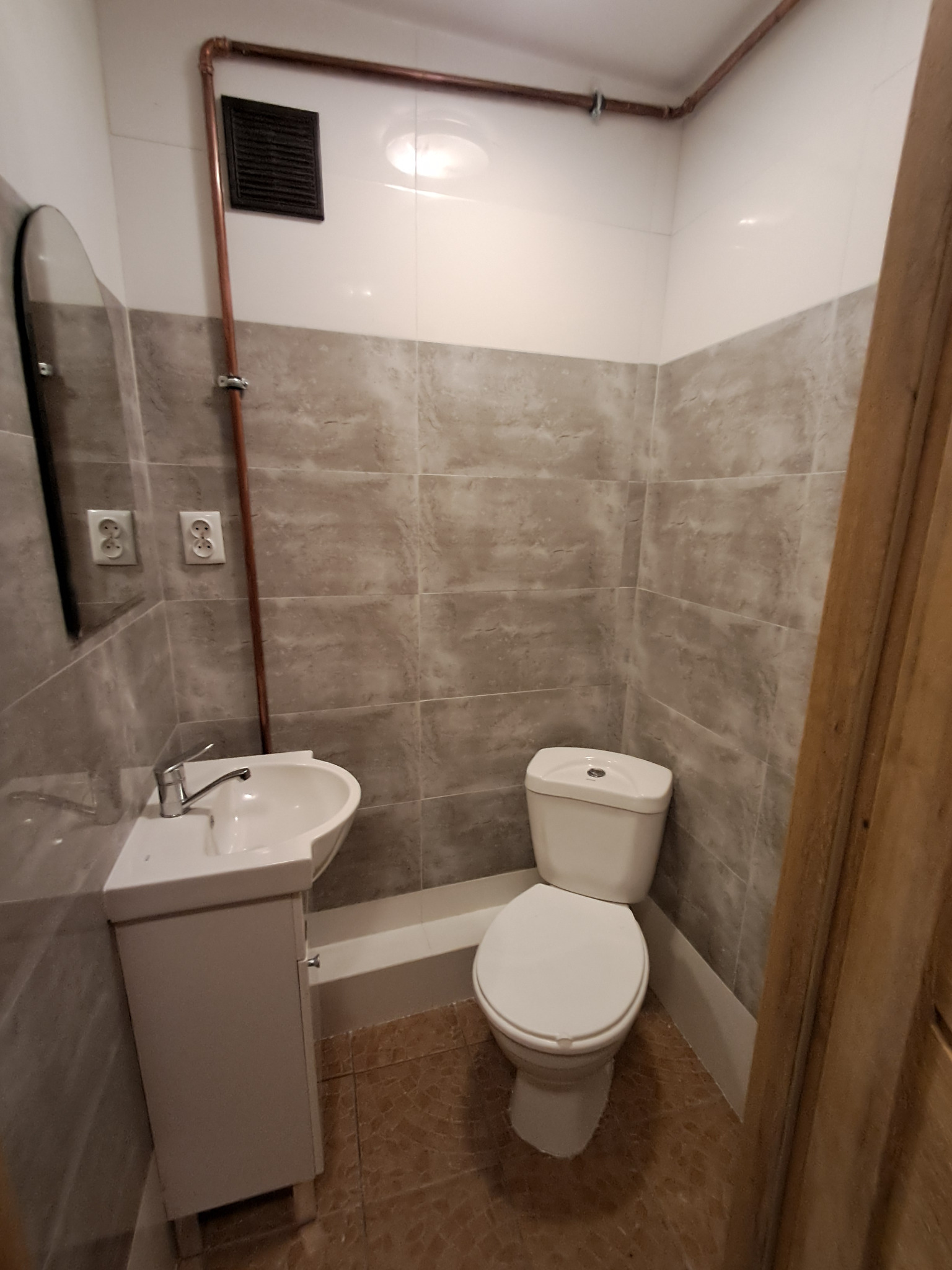 Wykończona łazienka z szarymi płytkami imitującymi kamień, białą umywalką z szafką, toaletą i miedzianą rurą biegnącą wzdłuż sufitu.