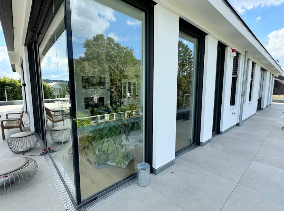 Nowoczesny taras z przesuwnymi drzwiami aluminiowymi z czarnymi ramami, widok na ogród odbity w szybie, minimalistyczne meble ogrodowe na zewnątrz.