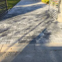 Nowoczesny podjazd z szarej kostki brukowej ułożonej w geometryczny wzór, prowadzący do białego domu z otwartą metalową bramą i zadbanym trawnikiem.