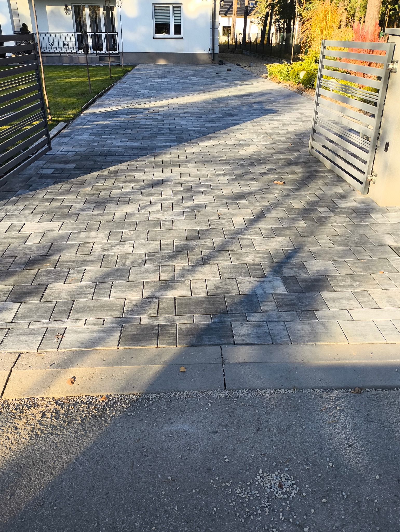 Nowoczesny podjazd z szarej kostki brukowej ułożonej w geometryczny wzór, prowadzący do białego domu z otwartą metalową bramą i zadbanym trawnikiem.