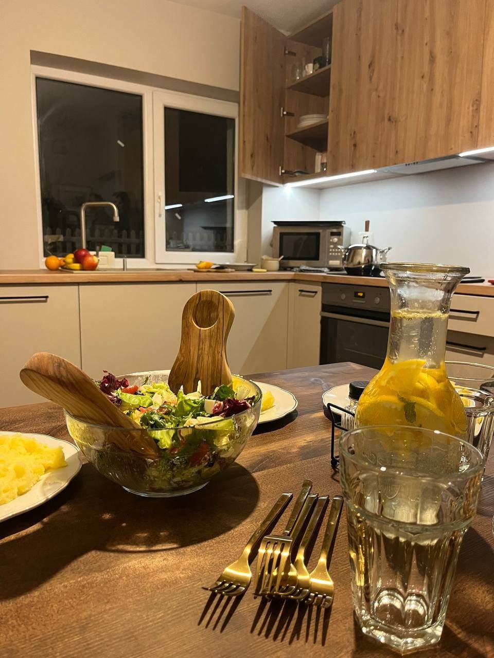 Nowoczesna, ciepła kuchnia z drewnianymi frontami szafek, oświetleniem LED pod szafkami wiszącymi i stołem z sałatką, puree ziemniaczanym i lemoniadą cytrynową, przygotowanymi do spożycia.