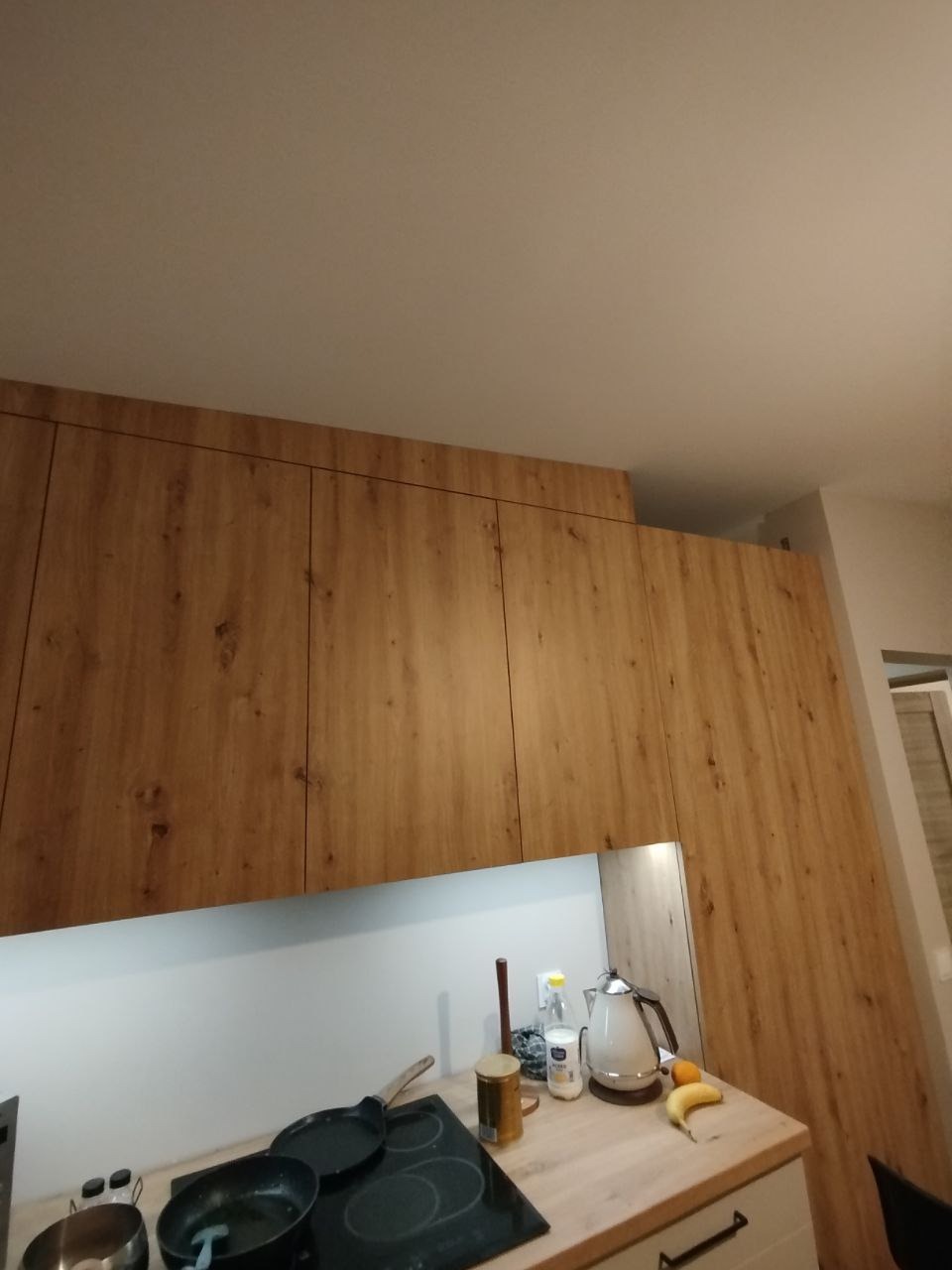 Wysokie szafki kuchenne z frontami w odcieniach drewna, sięgające sufitu, z widocznym blatem i częścią płyty grzewczej z czarnymi garnkami i czajnikiem.