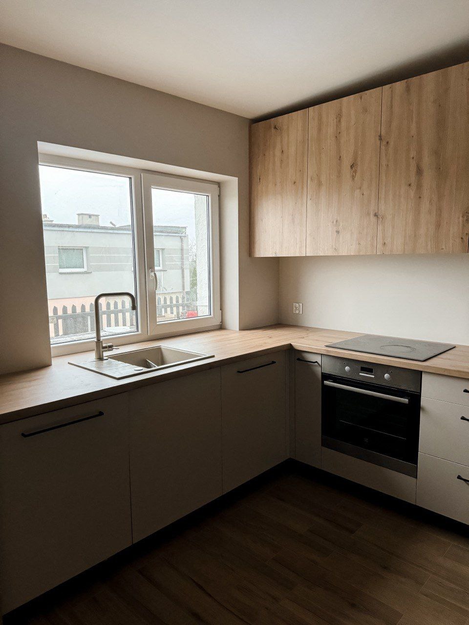 Jasna kuchnia z drewnianymi szafkami wiszącymi i blatem, z widokiem na szare budynki za oknem.