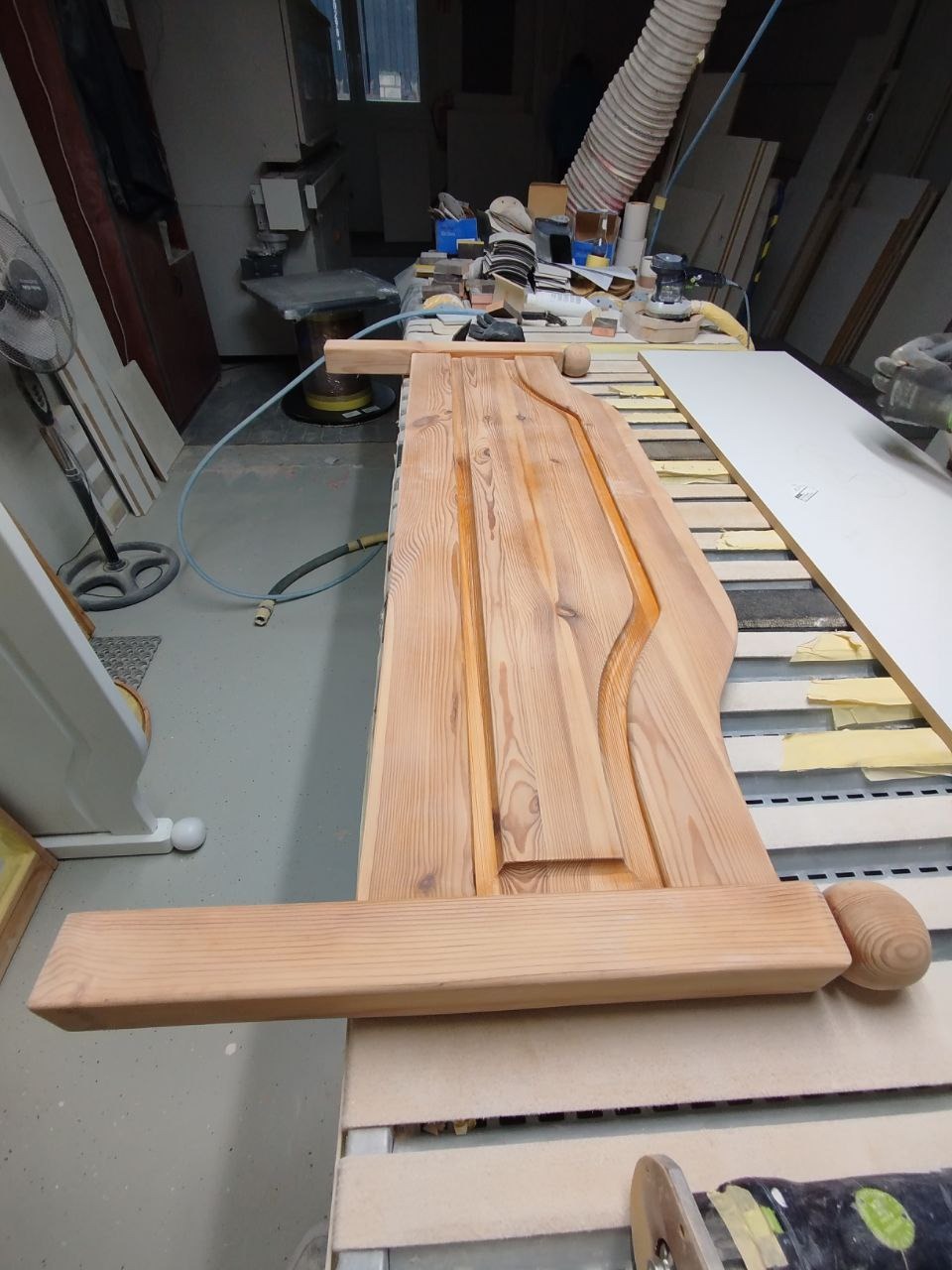 Drewniany, surowy front meblowy z ozdobnymi detalami, widoczny w warsztacie stolarskim, na stole roboczym obok narzędzi i innych elementów meblowych.