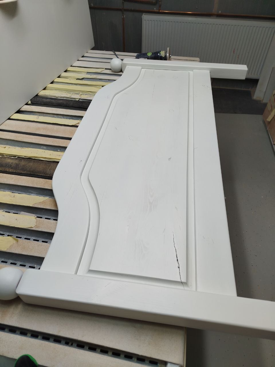 Biały, pomalowany front meblowy z dekoracyjnym frezem, leżący na stole warsztatowym; widoczne niedoskonałości malowania i pęknięcie.