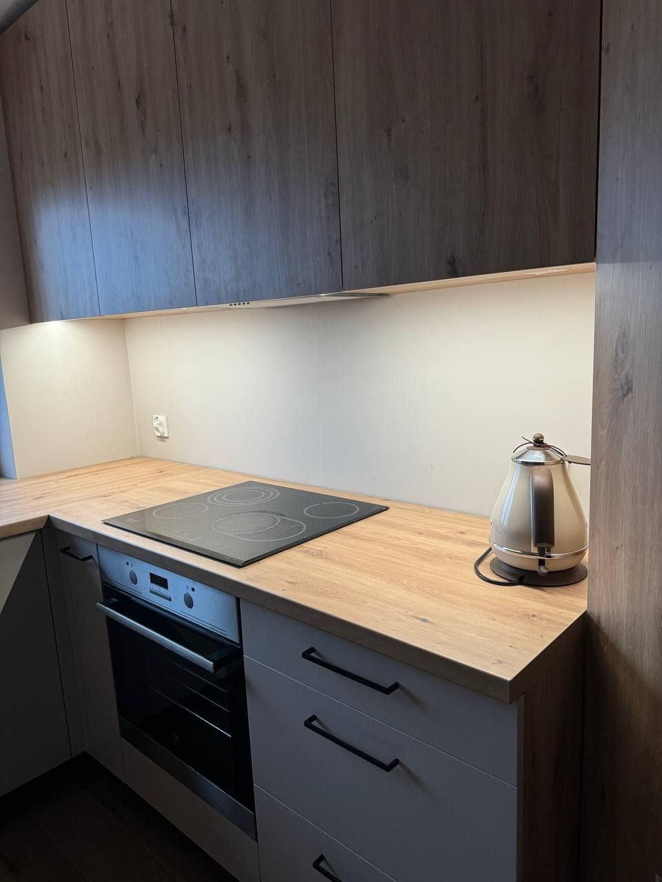Nowoczesna kuchnia z szarymi szafkami, drewnianym blatem, indukcyjną płytą grzewczą i beżowym czajnikiem elektrycznym, oświetlona listwą LED pod szafkami wiszącymi z ciemnego drewna.
