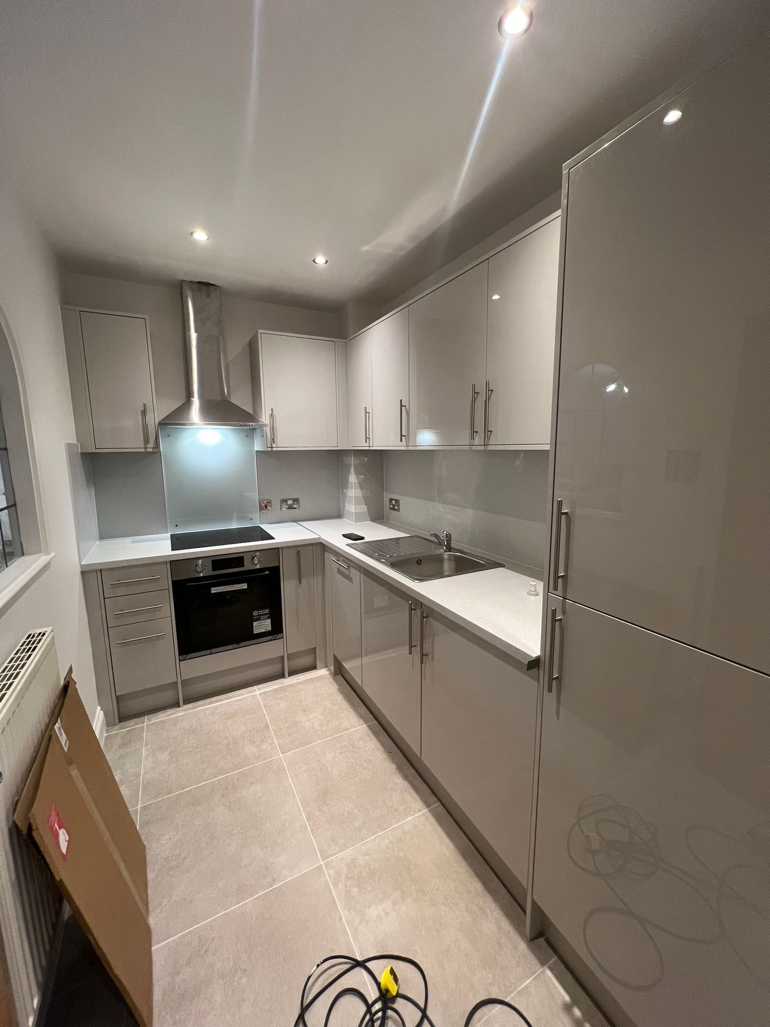 Nowoczesna kuchnia w odcieniach szarości z błyszczącymi frontami szafek, metalowym okapem i szklanym panelem nad blatem, widoczne kartony i kable na podłodze.