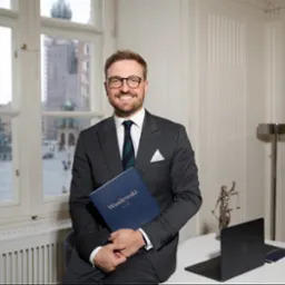 Uśmiechnięty mężczyzna w garniturze i okularach, siedzący na biurku w gabinecie, trzymający niebieski folder z napisem 'Wasilewski LEGAL', w tle widok na Rynek Główny w Krakowie, obok laptop...