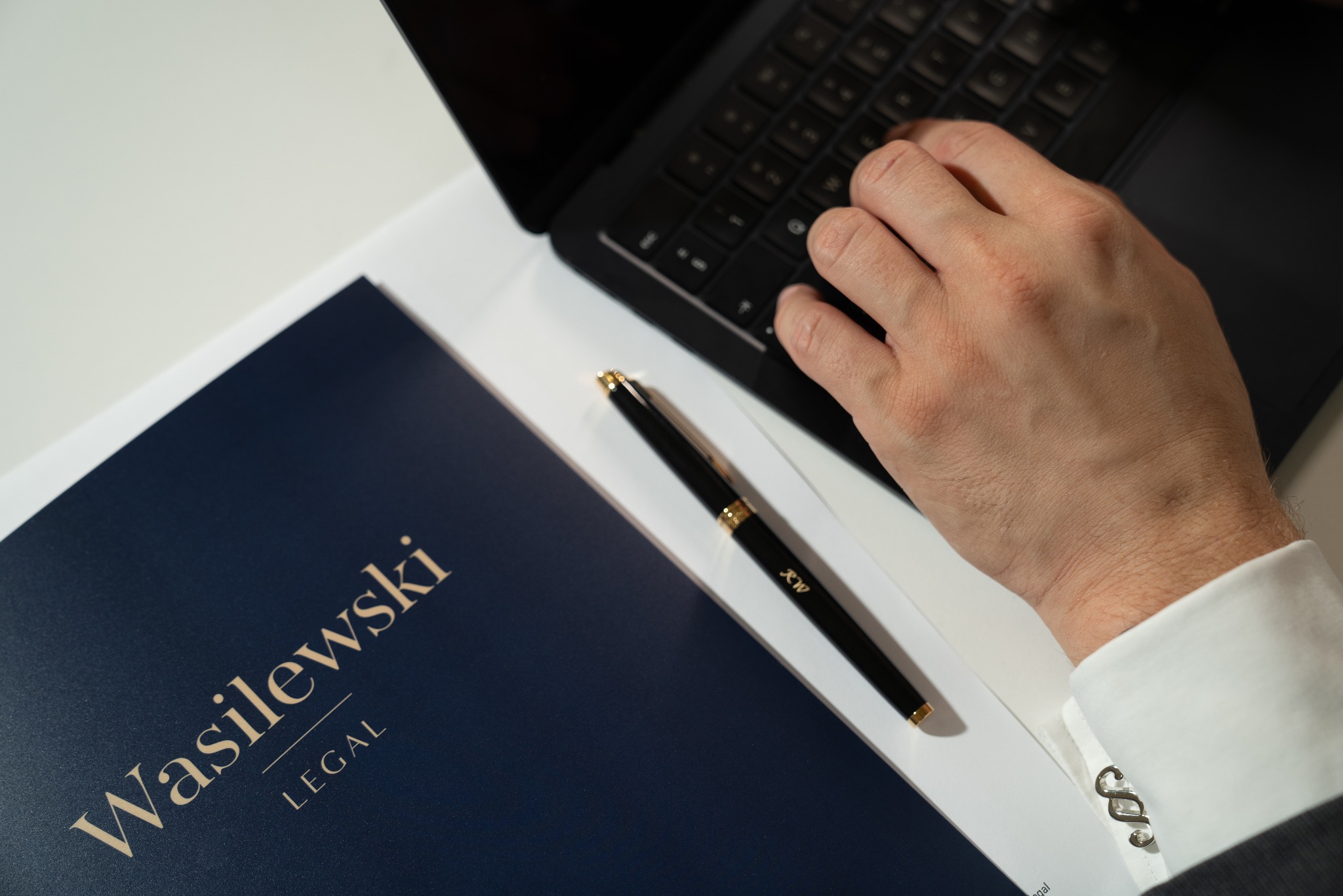 Dłoń mężczyzny piszącego na laptopie obok granatowej teczki z napisem Wasilewski Legal i czarnego pióra z inicjałami RW, widoczne spinki do mankietów w kształcie paragrafów.