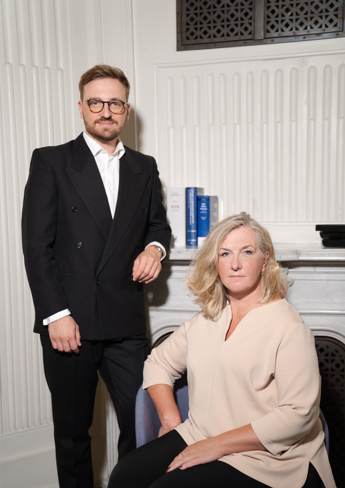 Portret dwojga prawników: mężczyzna w czarnym garniturze stoi obok kobiety w beżowej bluzce siedzącej na krześle, w tle biała ściana z grzejnikiem i książki