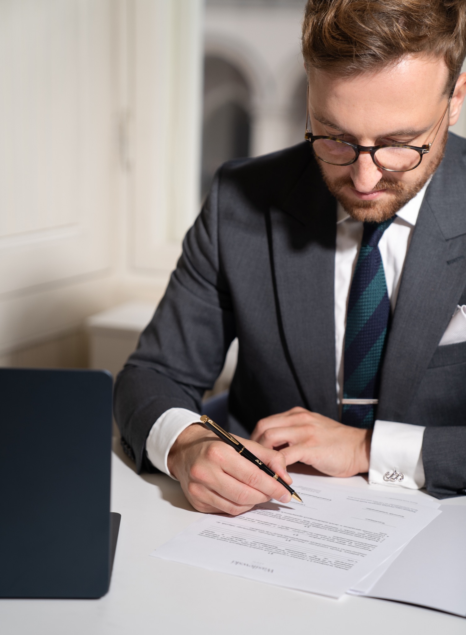 Mężczyzna w garniturze podpisuje dokument złotym piórem, widoczne mankiety koszuli i laptop w tle.