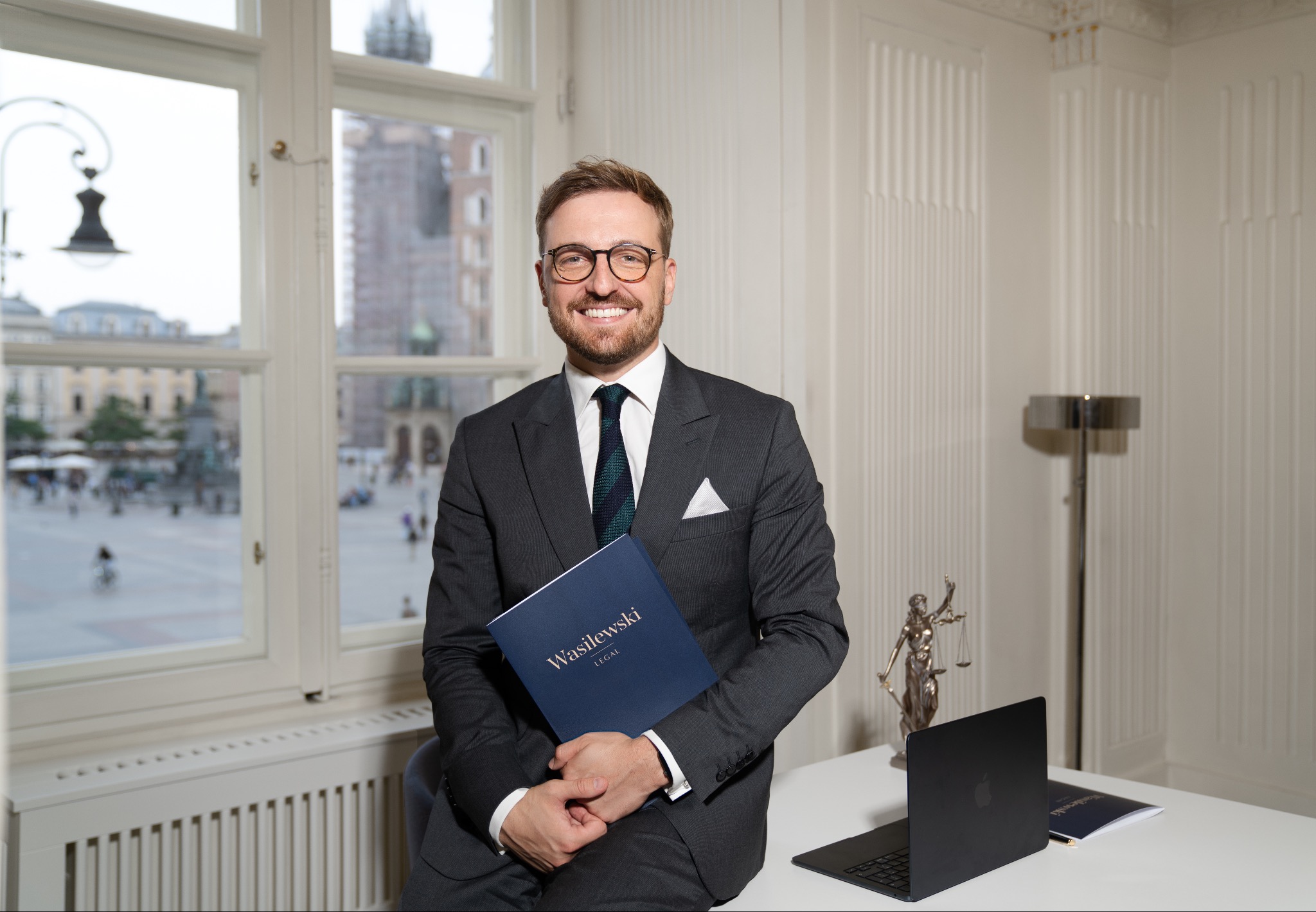 Uśmiechnięty mężczyzna w garniturze i okularach, siedzący na biurku w gabinecie, trzymający niebieski folder z napisem 'Wasilewski LEGAL', w tle widok na Rynek Główny w Krakowie, obok laptop...