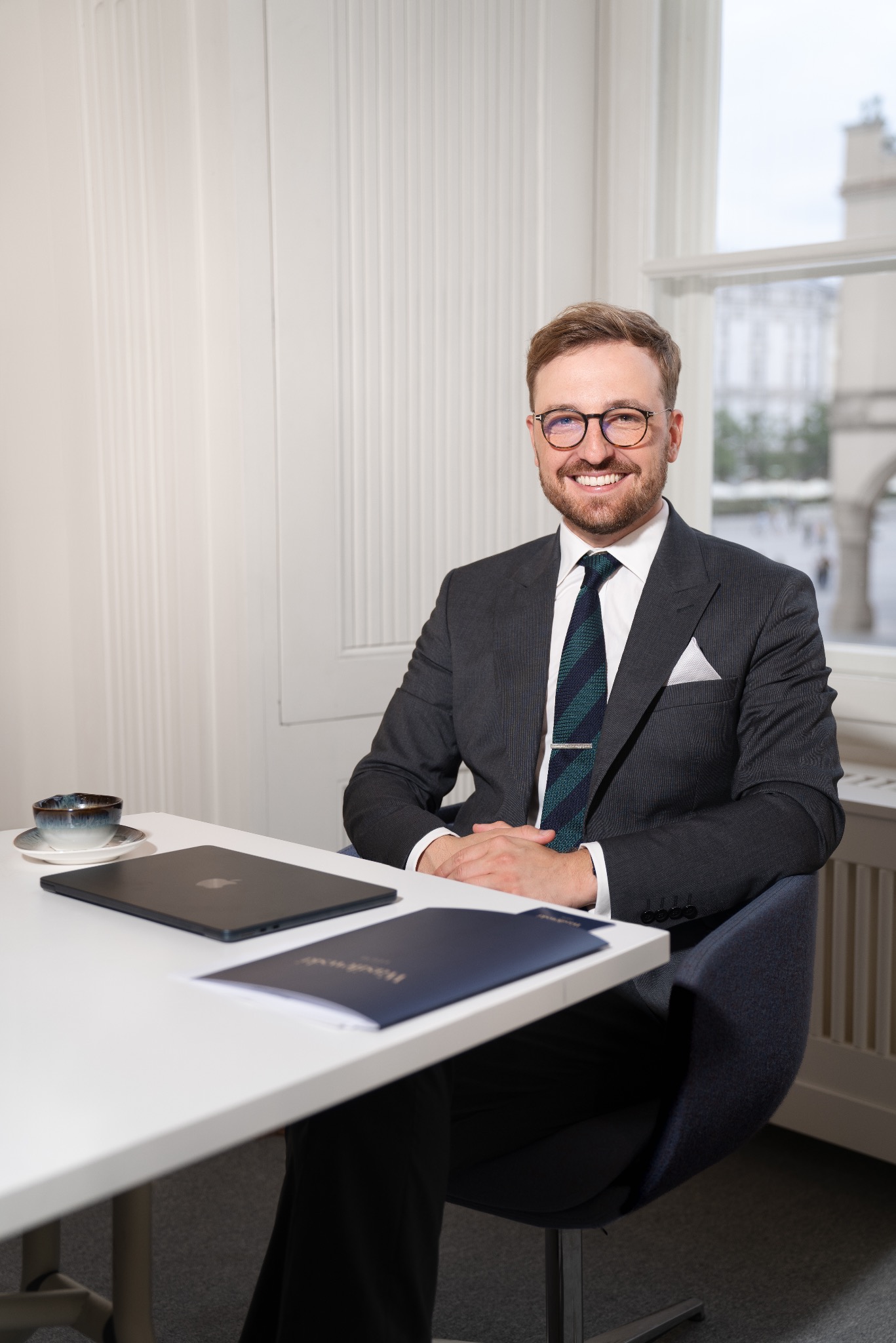 Uśmiechnięty mężczyzna w garniturze i okularach siedzi za biurkiem w jasnym biurze. Na biurku laptop, filiżanka i niebieska teczka.