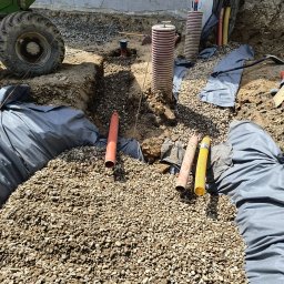IP INVEST - Wykonane wykopy z ułożonymi rurami i zasypane żwirem, zabezpieczone geowłókniną, widoczny fragment maszyny budowlanej.