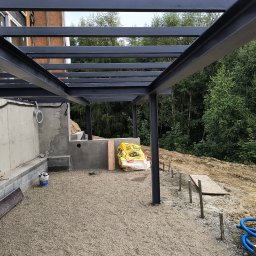 IP INVEST - Konstrukcja stalowa z profili, prawdopodobnie zadaszenia, w trakcie montażu, widoczne fundamenty i przygotowane podłoże z kruszywa, w tle drzewa.
