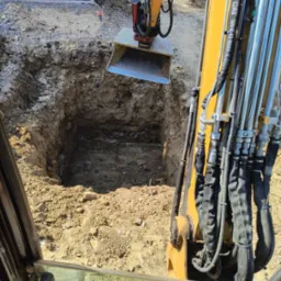 Żółta koparka z łyżką pogłębia wykop w ziemi, widok z kabiny operatora, w tle fundamenty budynku.