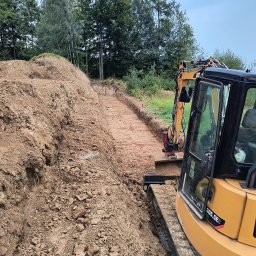 IP INVEST - Żółta koparka gąsienicowa CAT 305.5E2 w trakcie prac ziemnych, widoczny wykop w gruncie, w tle zieleń drzew