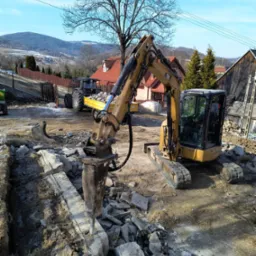 Żółty minikoparka z młotem pneumatycznym kruszy betonowy fundament, w tle traktor z przyczepą i zabudowania wiejskie.