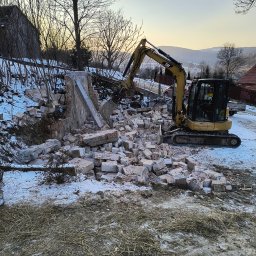 IP INVEST - Żółta minikoparka CAT w trakcie rozbiórki murowanej konstrukcji na zaśnieżonym terenie, z widocznymi górami w tle i zachodzącym słońcem.