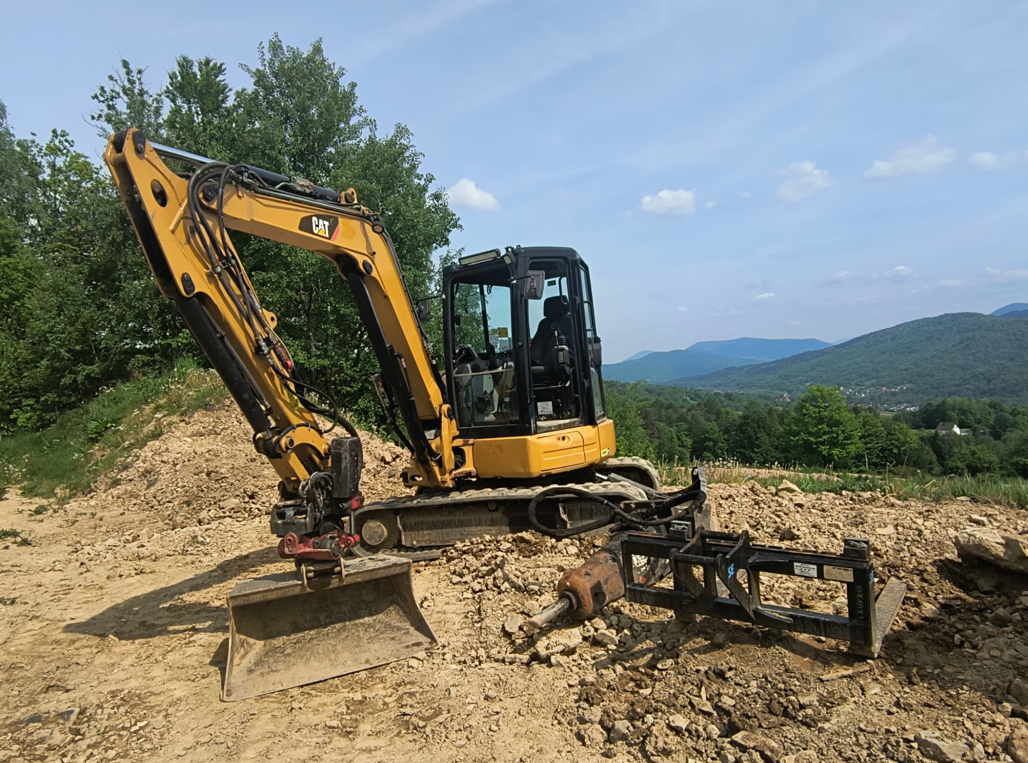 Żółta koparka CAT na nierównym terenie z kamieniami, widok na zalesione wzgórza i błękitne niebo w tle.