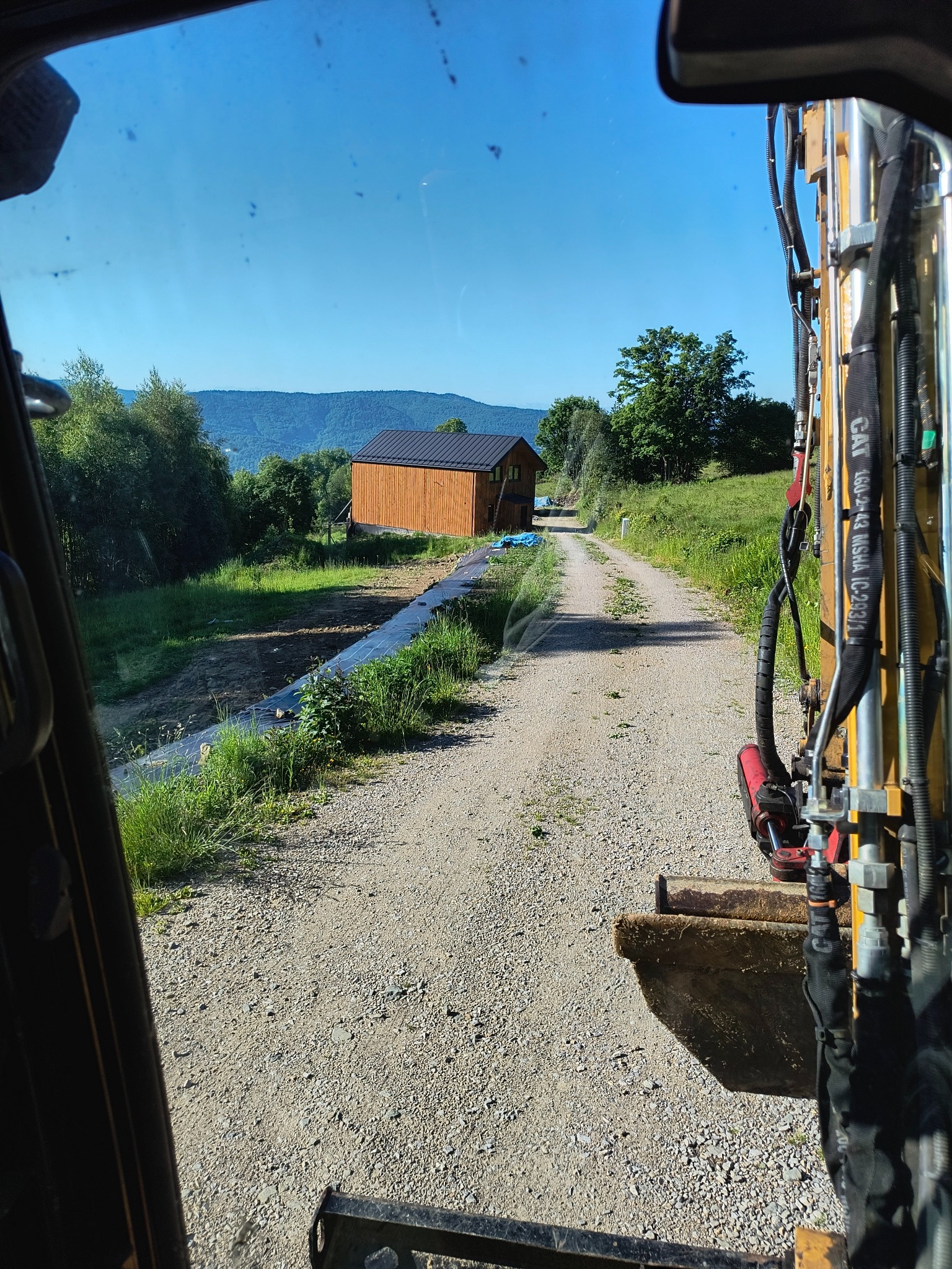 Widok z kabiny koparki na szutrową drogę prowadzącą do drewnianego budynku gospodarczego na tle zalesionych wzgórz.