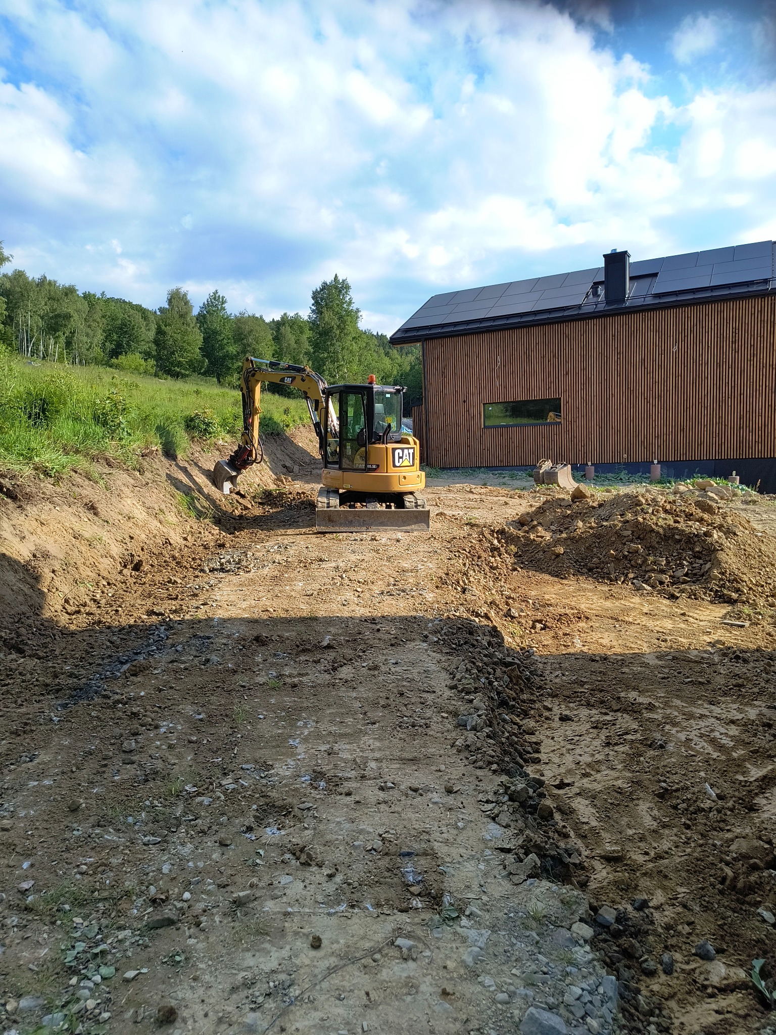 Żółta mini koparka CAT podczas niwelacji terenu pod budowę domu w Tokarni, widoczne wykopane rowy i pryzma ziemi, w tle nowoczesny dom z panelami słonecznymi na dachu.