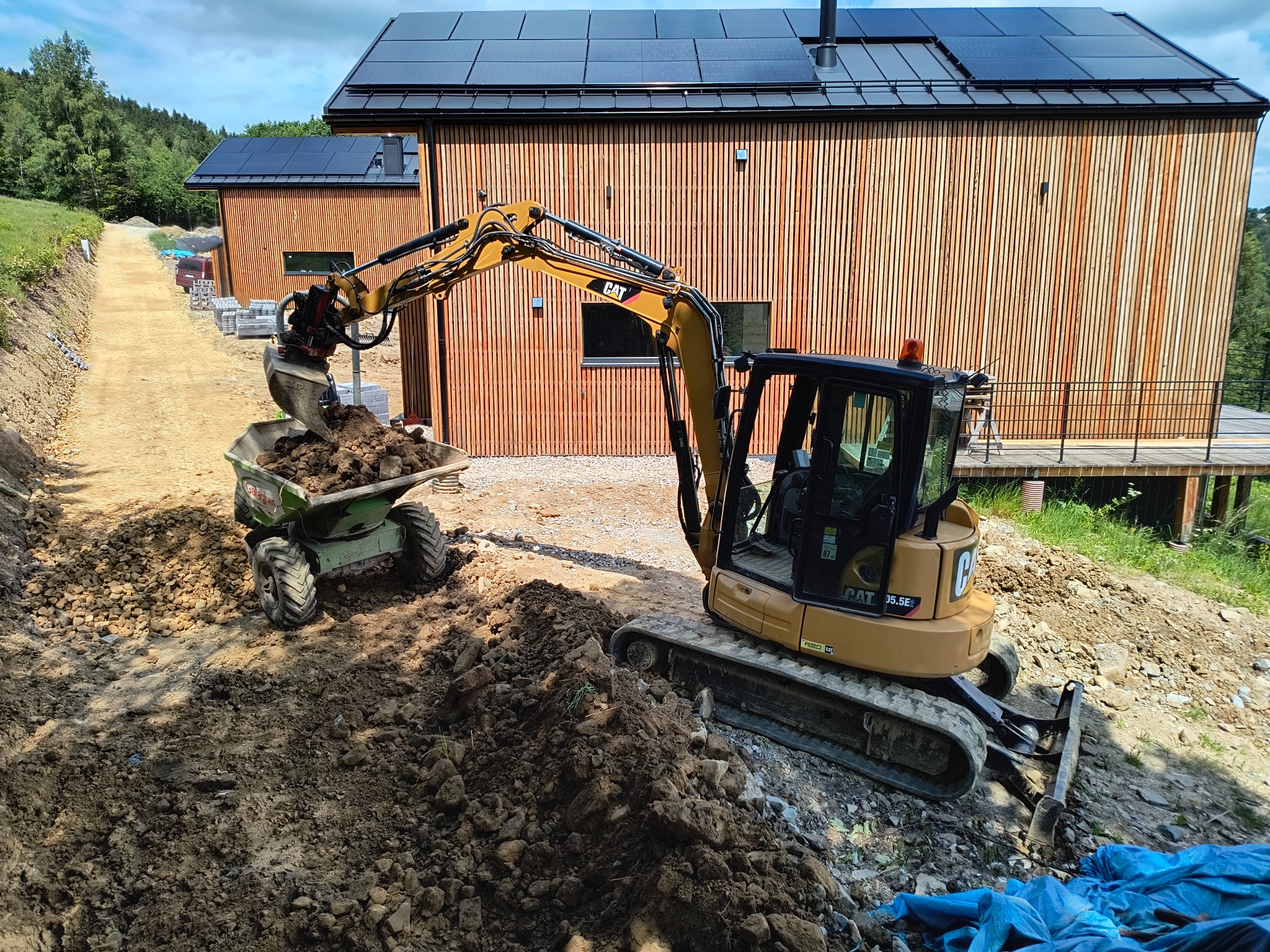 Żółta minikoparka CAT 30.5E nabiera ziemię do zielonej taczki na dwóch kołach, w tle budynek z drewnianą elewacją i panelami słonecznymi na dachu oraz droga gruntowa.