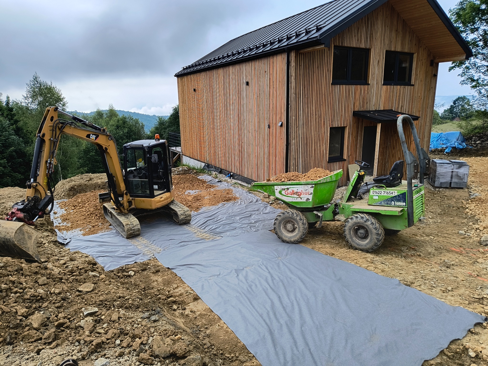 Żółta mini koparka CAT i zielona wywrotka Gallagher na placu budowy z nowym domem o drewnianej elewacji w tle, teren zabezpieczony szarą geowłókniną.