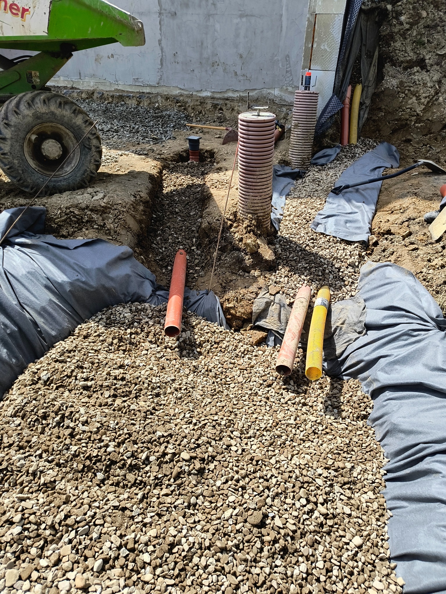 Wykonane wykopy z ułożonymi rurami i zasypane żwirem, zabezpieczone geowłókniną, widoczny fragment maszyny budowlanej.