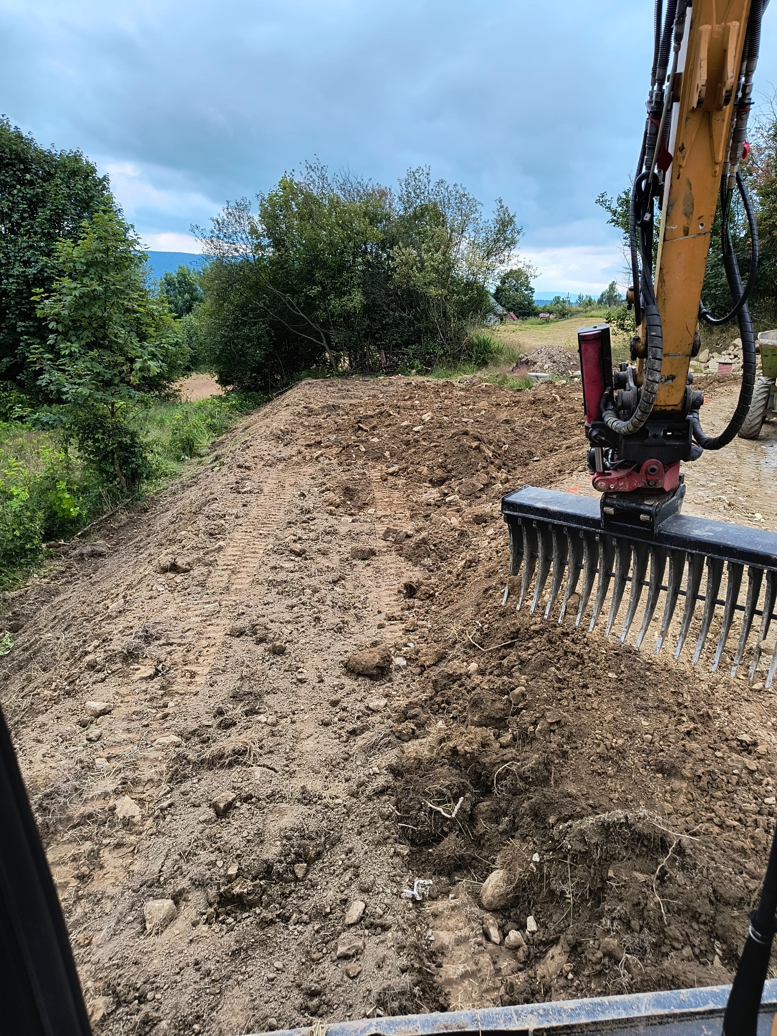 Żółta koparka z nasadką w kształcie grabi wyrównuje teren, w tle widoczne drzewa i zarys gór przy pochmurnym niebie.