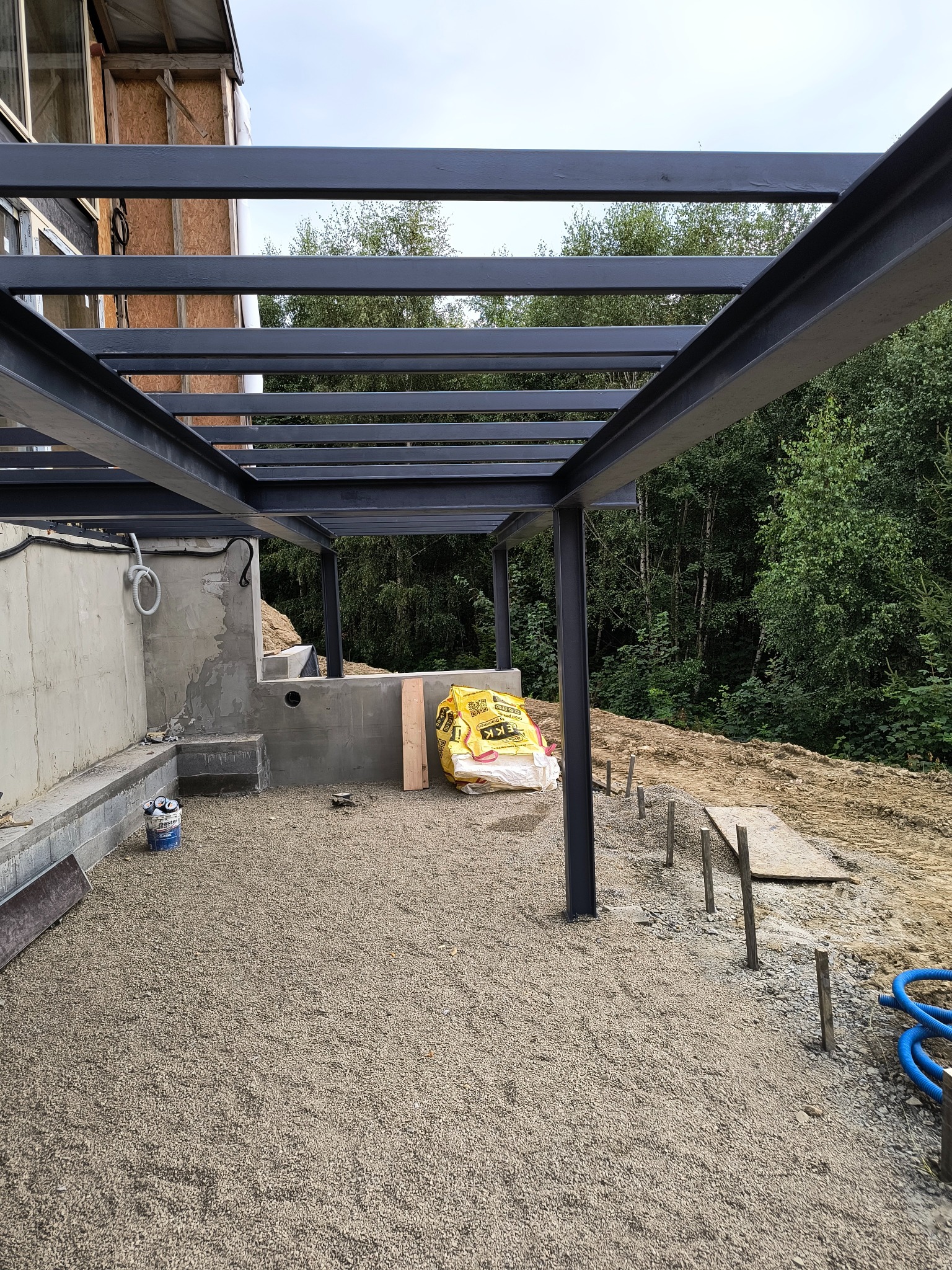 Konstrukcja stalowa z profili, prawdopodobnie zadaszenia, w trakcie montażu, widoczne fundamenty i przygotowane podłoże z kruszywa, w tle drzewa.