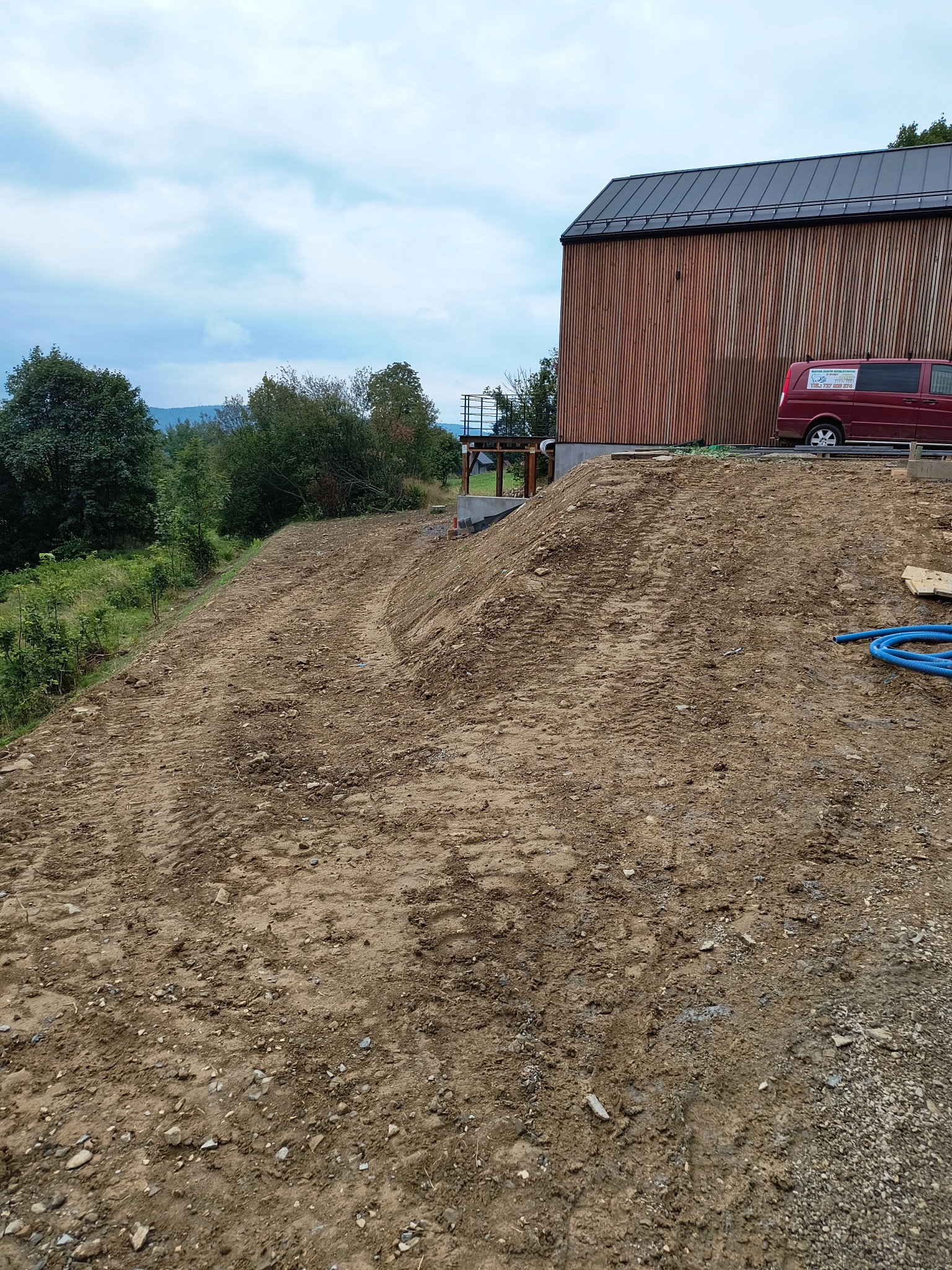 Niwelacja terenu pod budowę domu z drewnianą elewacją, widoczne ślady opon i niebieski wąż, w tle zieleń i góry.